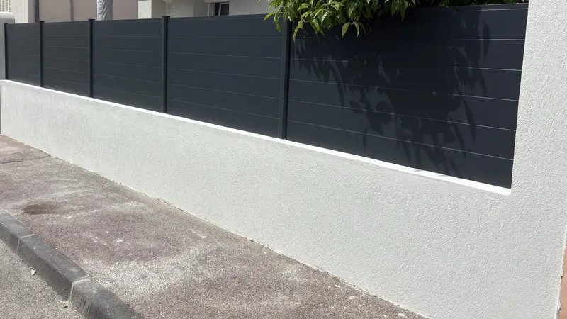Clôture en lames aluminium anthracite posée sur un muret enduit clair, le long d'un trottoir