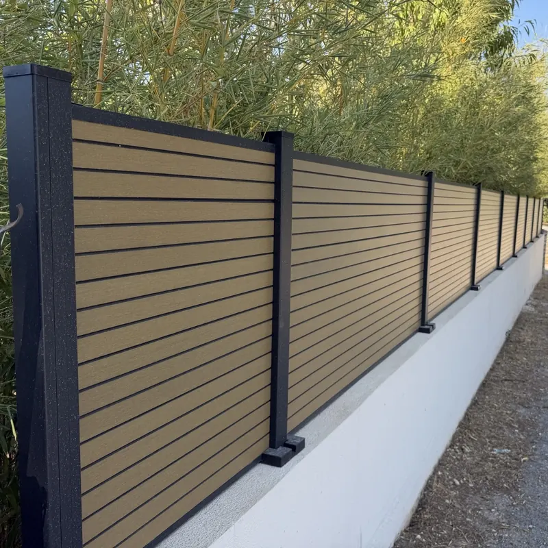 Clôture en lames composite ton bois clair entre poteaux aluminium noirs, sur un muret enduit blanc