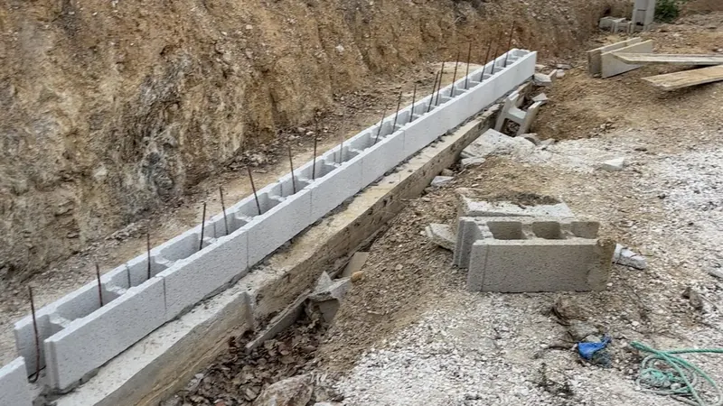Premier rang de blocs béton d'un muret posé sur sa semelle, fers en attente dans les alvéoles, au pied d'un talus