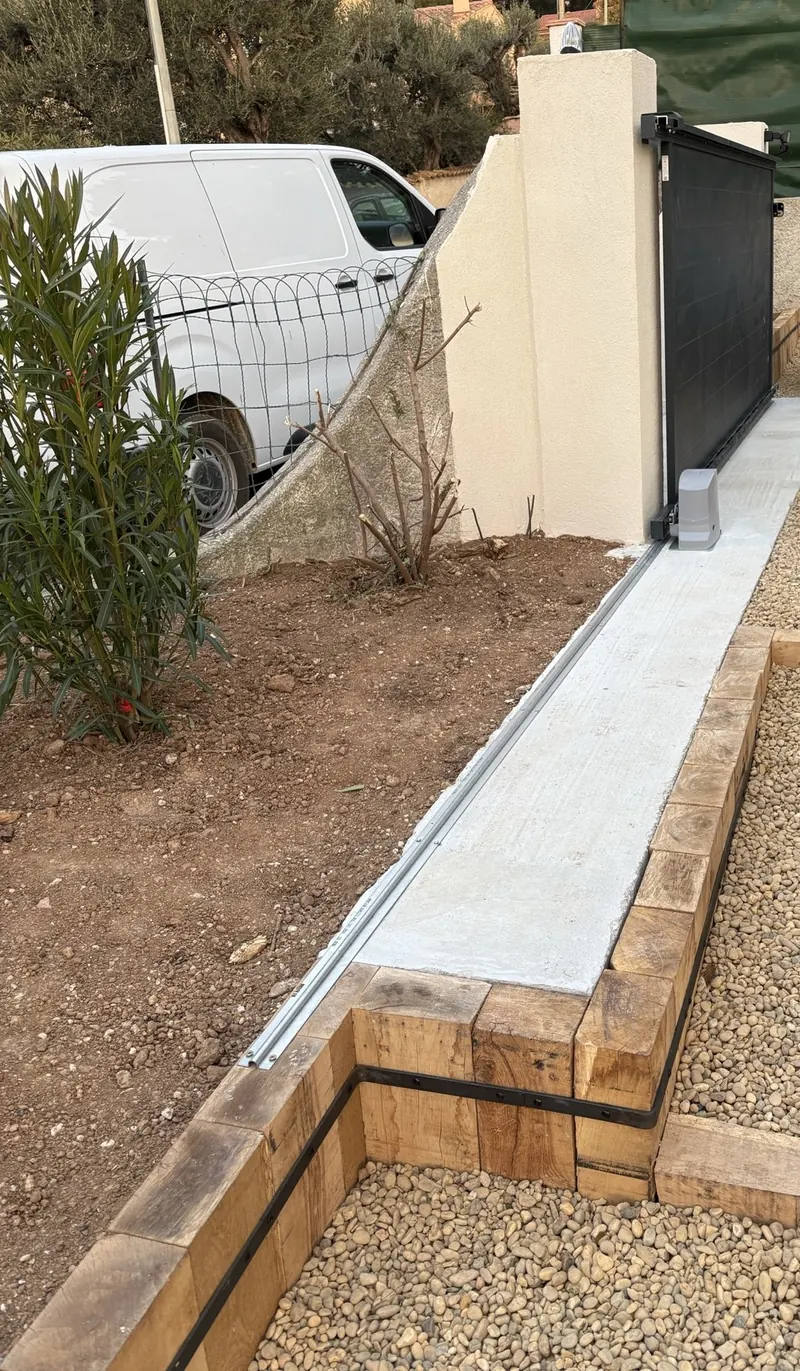 Portail coulissant anthracite motorisé, rail fixé sur une longrine béton le long d'une allée bordée de traverses