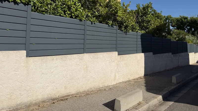 Clôture en lames aluminium anthracite posée en redans sur un muret enduit qui suit la pente du trottoir