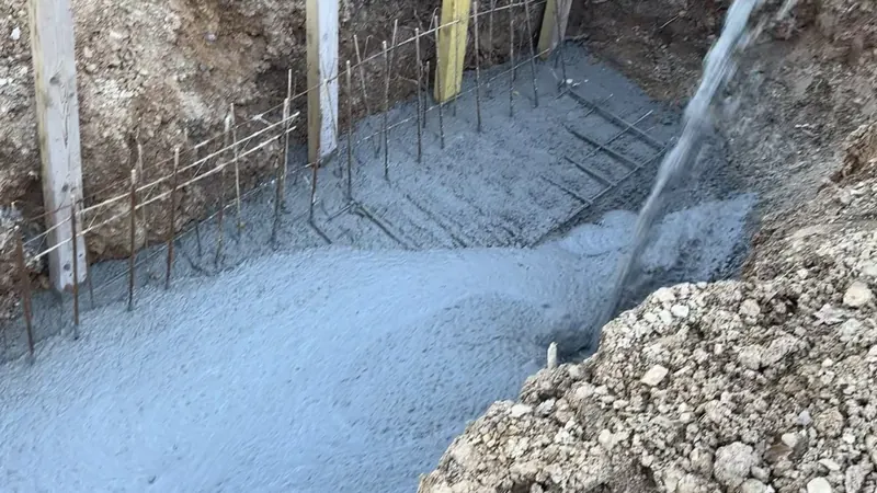 Béton coulé dans la fouille ferraillée de la fondation d'un muret, fers en attente et piquets d'implantation